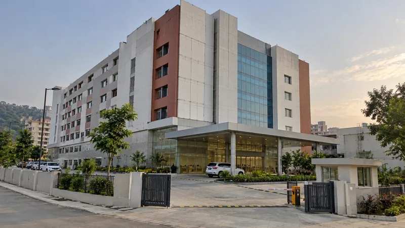 Illustrative render of a multi-storey hospital building with white and terracotta panels and a glass tower, a covered car porch and glazed entrance, a gated drive with a boom barrier and security cabin, and green hills behind