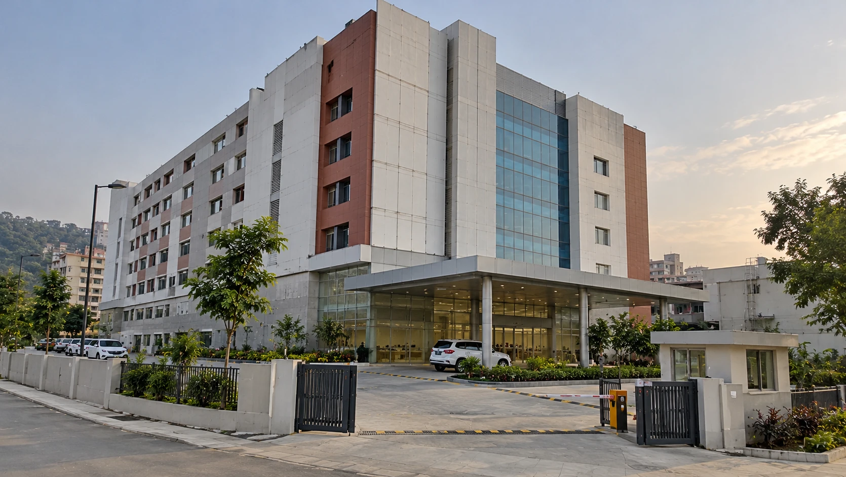 Illustrative render of a multi-storey hospital building with white and terracotta panels and a glass tower, a covered car porch and glazed entrance, a gated drive with a boom barrier and security cabin, and green hills behind